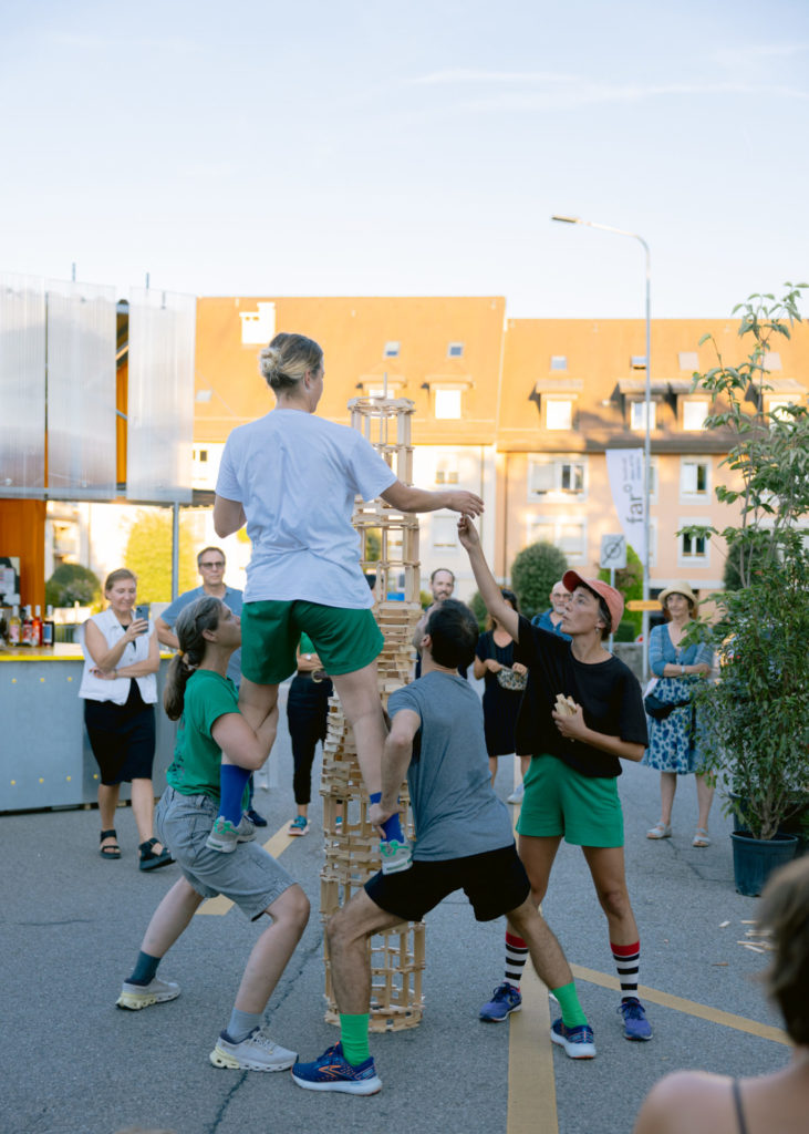 The artists of the Dénominateurs Communs collective construct a high tower with kaplas. One of the artists stands on top of two other artists' legs.