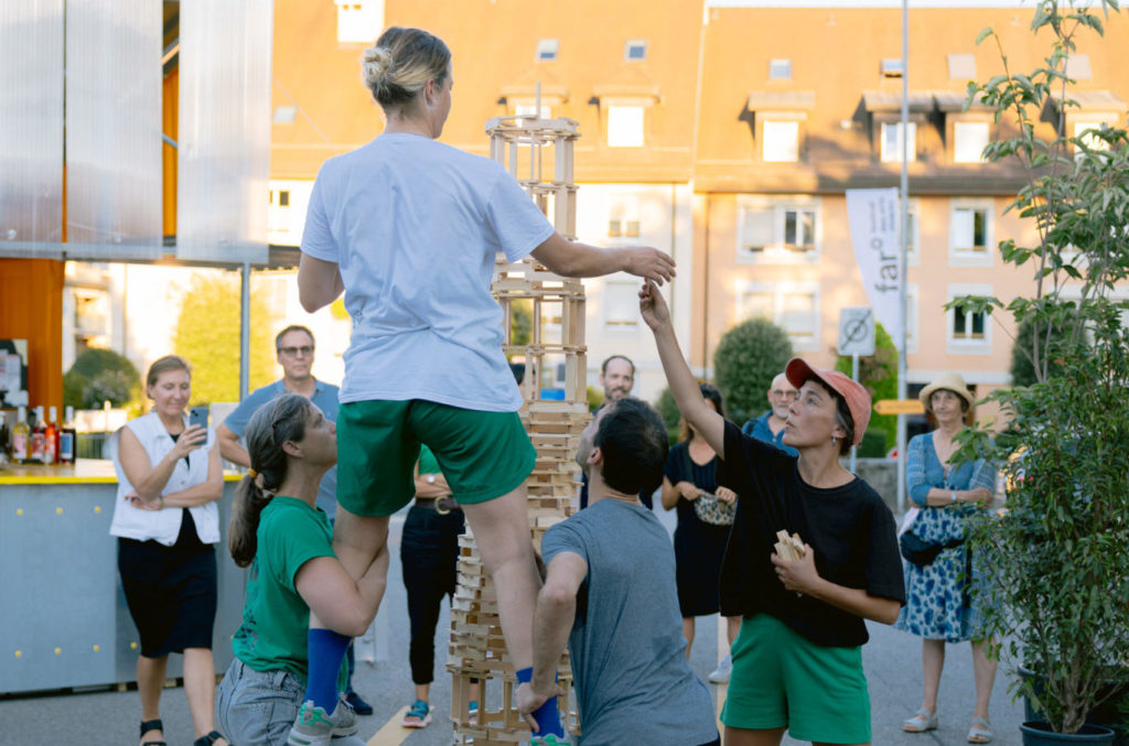 Quatre personnes du collectif Dénominateurs Communs construisent une grande tour avec des kaplas. Une des artistes se tient surélevée sur les jambes de deux autres artistes.