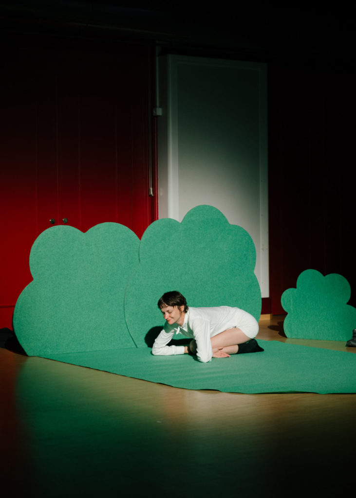 The comedian Marion Thomas sits on a dark green moquet that takes the form of bushes, in a sheep-like manner.