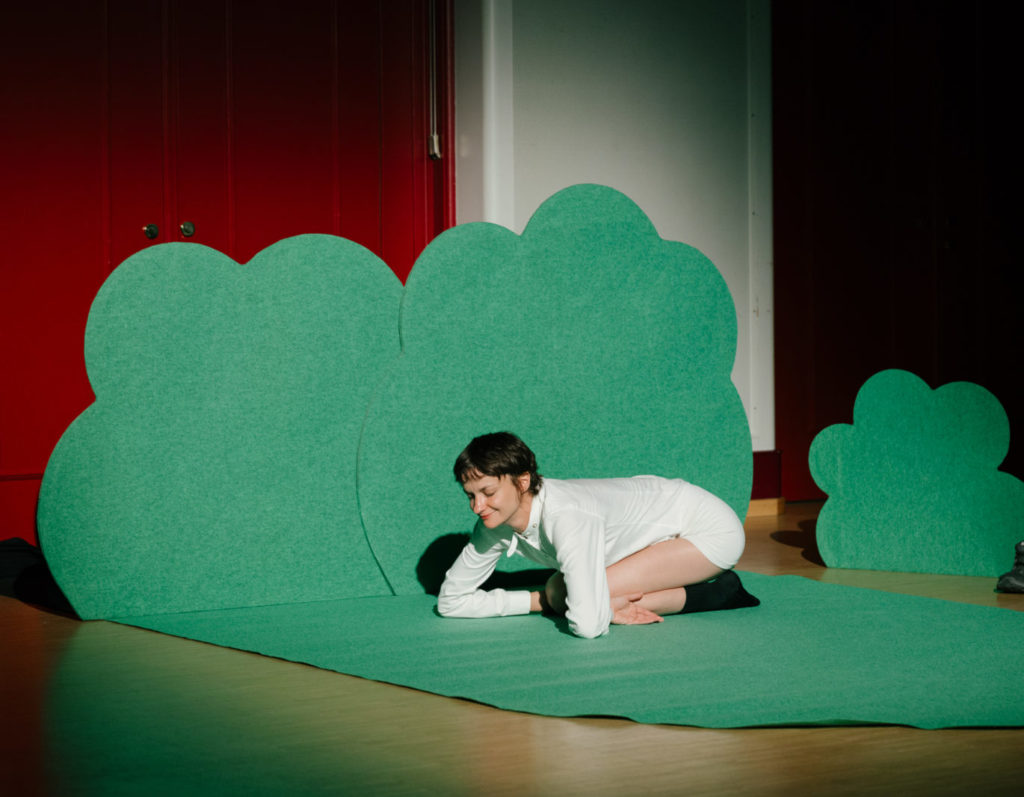 La comédienne Marion Thomas se tient comme un mouton, par terre, sur une moquette verte en forme de buisson.