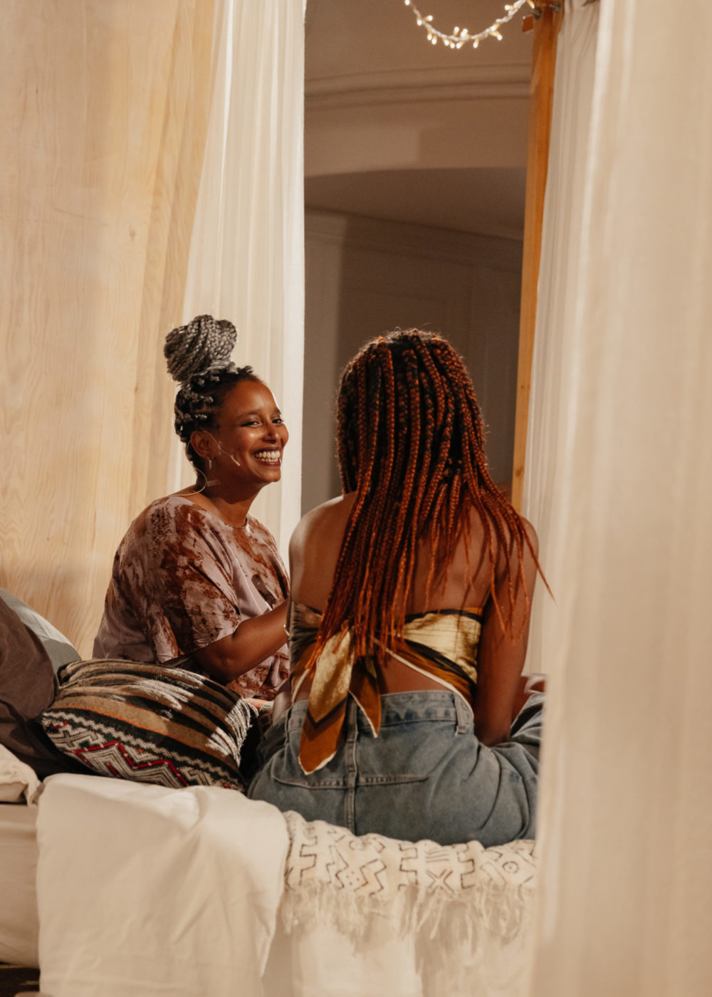 L'artiste Lucile Saada Choquet se tient de face sur un lit et regarde une personne à sa droite, venue pour participer à sa performance et parler avec l'artiste de son vécu en tant que personne racisée.