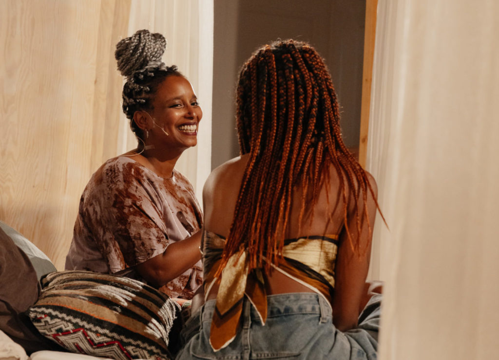 L'artiste Lucile Saada Choquet se tient de face sur un lit et regarde une personne à sa droite, venue pour participer à sa performance et parler avec l'artiste de son vécu en tant que personne racisée.