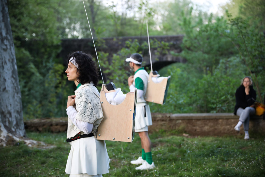 La scène a lieu dehors, dans une étendue d'herbe. On voit deux personnes tenir des sacs construits en bois avec des antennes radio qui dépassent.