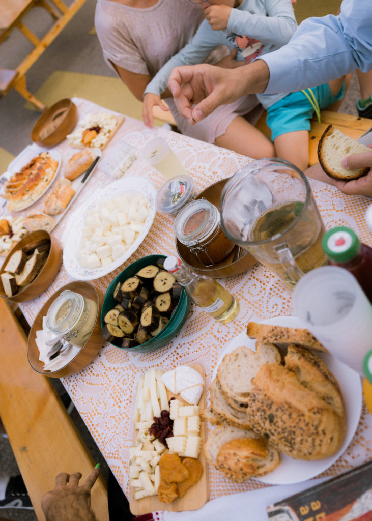 On a table are presented different dishes and beverages. There is bread, cheese, plantain bananas and bread for example. Hands are approaching the table to grasp something to eat.