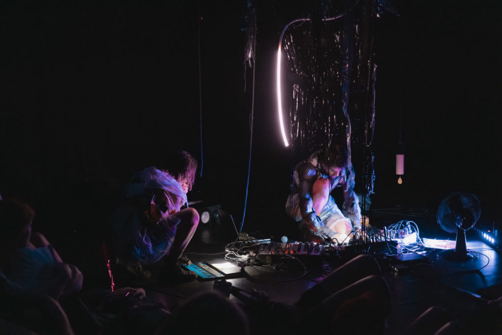 On voit l'artiste Francesca Sproccati à gauche par terre, qui crée des sons sur un grand synthé. À sa droite, un musicien fait de même. Il fait sombre mais il y a des néons violets qui éclairent la salle.