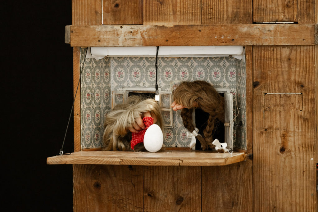 À travers une ouverture d'une boite en bois, on aperçoit deux mains. Elles sont recouvertes de perruques et se font face comme des personnages. Elles sortent de deux petites portes et observent un oeuf qui est au centre.