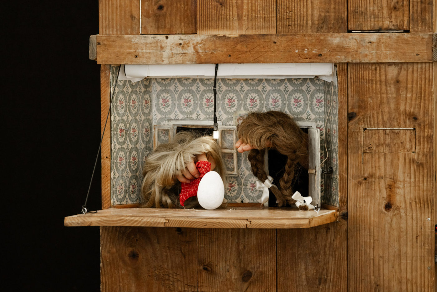 À travers une ouverture d'une boite en bois, on aperçoit deux mains. Elles sont recouvertes de perruques et se font face comme des personnages. Elles sortent de deux petites portes et observent un oeuf qui est au centre.