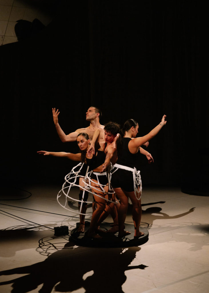 The four waltz dancers stand on a little and circular black platform and they form a circle. The bodies tries to escape the circle. We notice this through the arms that point toward the top of the room and the gazes in different directions. The costumes are mostly black but some parts are white.