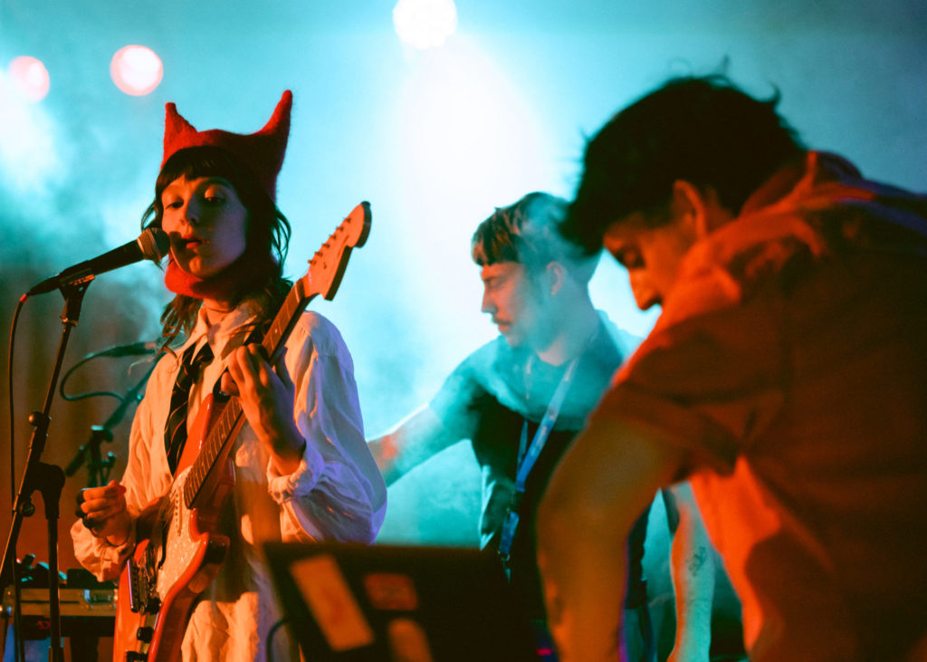 À gauche se trouve la chanteuse de 24 KIARA. Elle porte un bonnet rouge avec deux pics, comme des oreilles de chat. Derrière elle on aperçoit les deux autres musiciens, un au clavier et un à la guitare.