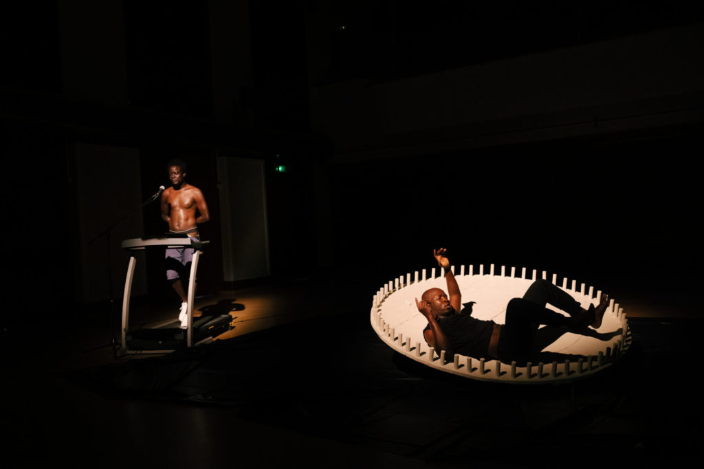 Les deux artistes se trouvent sur une scène. Mbaye Diop sur une machine de sport qui le fait marcher sur place. Il a les bras dans le dos et parle dans un micro. L'autre est sur le dos contre une paroi en bois circulaire qui le supporte. Il a les bras en l'air.