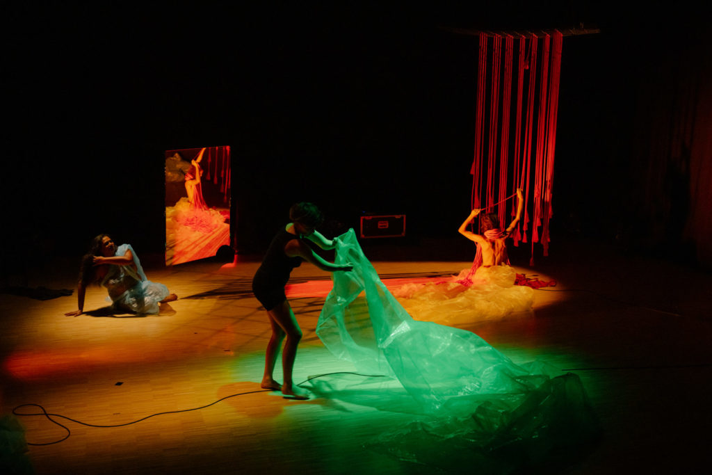 Les trois artistes occupent trois espaces différents de la scène. À gauche par terre, Living Smile Vidya est assise et remonte ses cheveux, dans son costume de plastique bleu. Au centre, Meret Landolt s'empare d'un plastique vert avec ses bras courts. À droite, Nina Langensand est nue, de dos, et s'enroule des cordes rouges qui la couvrent progressivement.