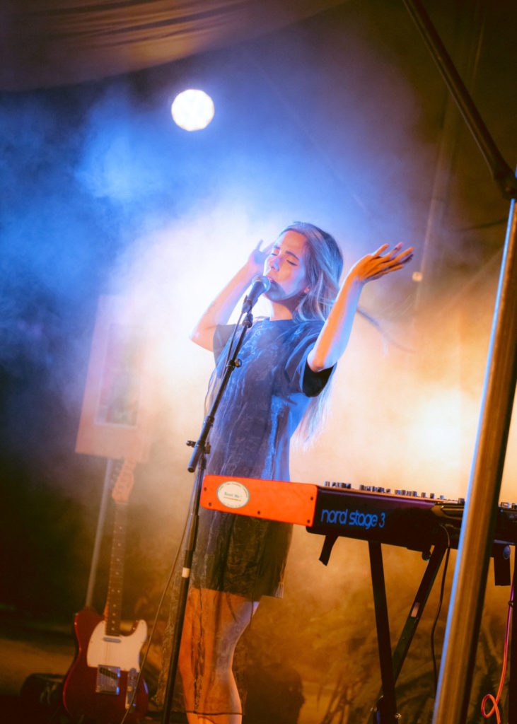 La chanteuse Verde Prato se tient debout sur la scène. Elle se tient derrière son clavier rouge. Ses avant-bras sont en l'air et elle a les yeux fermés.
