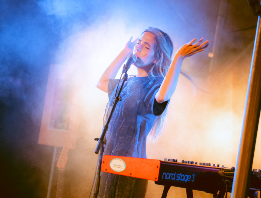 La chanteuse Verde Prato se tient debout sur la scène. Elle se tient derrière son clavier rouge. Ses avant-bras sont levés en l'air et elle a les yeux fermés.