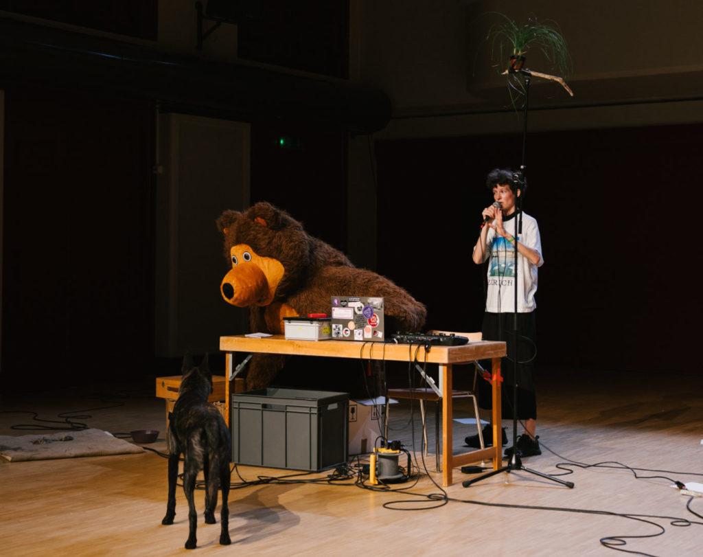 La comédienne Elise Perrin se tient sur scène. Elle et son chien regardent un ours en peluche gonflable sur la table. Il y a aussi une plante qui est placée tout en haut d'une tige de micro.