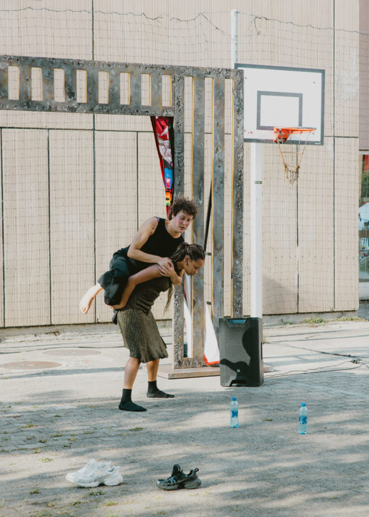 Une des deux danseuses soutient l'autre en la portant sur son dos. Elles se trouvent à côté d'un terrain de basket et on aperçoit une enceinte derrière elles.