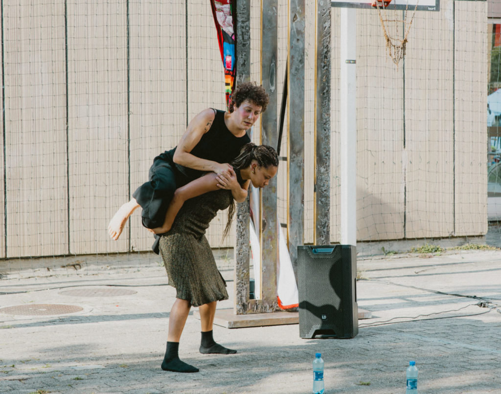 One of the two dancers supports the other on her back. They have removed their shoes. The performance occurs next to a basketball court et we notice a speaker in the back.