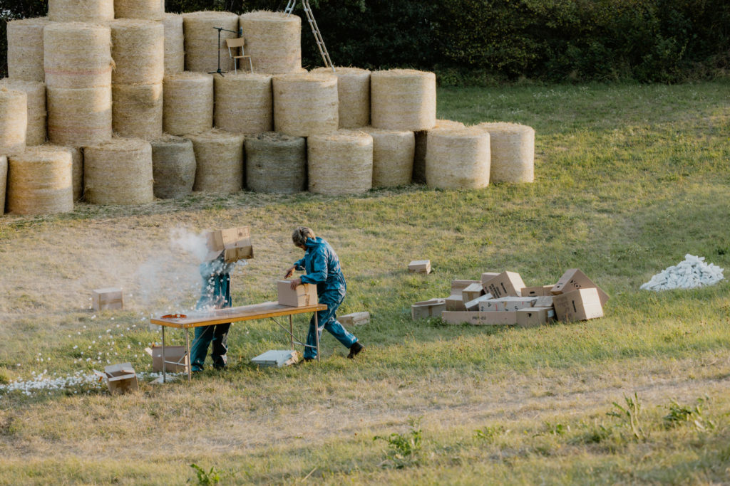 Deux comédiens se tiennent debout dans un champ. Il y a un montage de bottes de foin en arrière plan. L'un des deux ouvre un colis tandis que l'autre verse le contenu d'un autre colis sur sa tête. Il en ressort de la poussière et des morceaux de polystyrène.