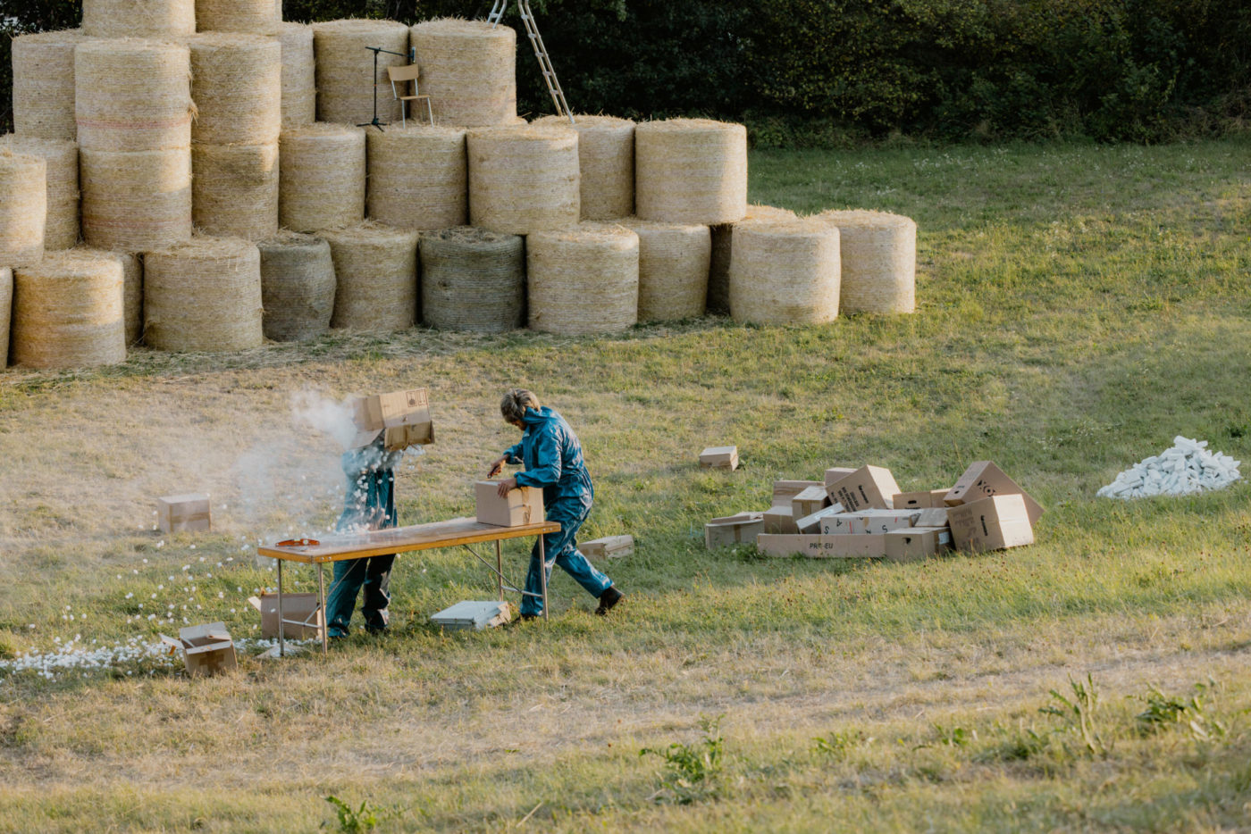 Deux comédiens se tiennent debout dans un champ. Il y a un montage de bottes de foin en arrière plan. L'un des deux ouvre un colis tandis que l'autre verse le contenu d'un autre colis sur sa tête. Il en ressort de la poussière et des morceaux de polystyrène.