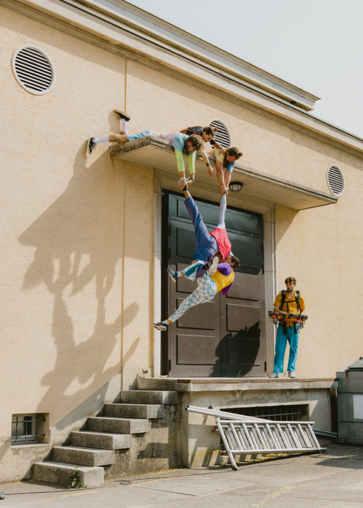 Three circus artists lie on a high point on a roof. They support another circus artist with their hands by grasping one of her legs and one of her hands. The suspended artist also supports another one. A musician stands on the right.