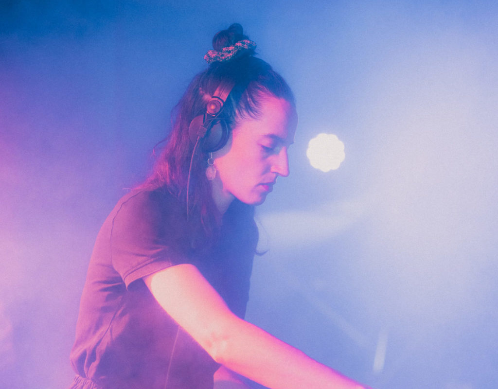 On voit la DJette awka mixer des sons. La main droite est visible et elle porte un casque. Elle baigne dans des lumières violettes, dans une légère fumée artificielle.