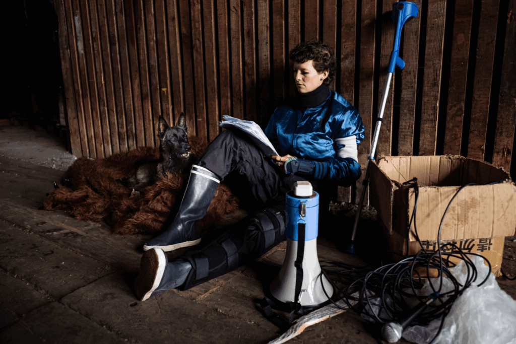 Elise Perrin est assise contre un mur en bois. Elle porte des bottes cirées pour la pluie. Elle a une jambe étendue, qui porte une attelle. Elle est en train de lire un texte. Il y a un carton à côté d'elle, un chien, un mégaphone et une béquille.