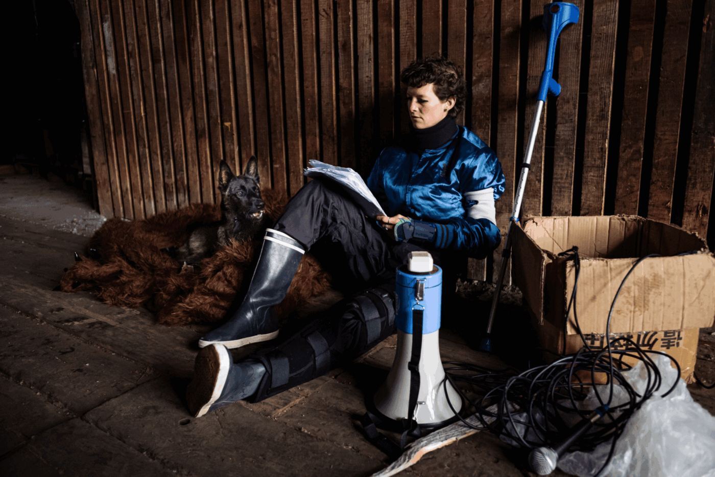 Elise Perrin est assise contre un mur en bois. Elle porte des bottes cirées pour la pluie. Elle a une jambe étendue, qui porte une attelle. Elle est en train de lire un texte. Il y a un carton à côté d'elle, un chien, un mégaphone et une béquille.