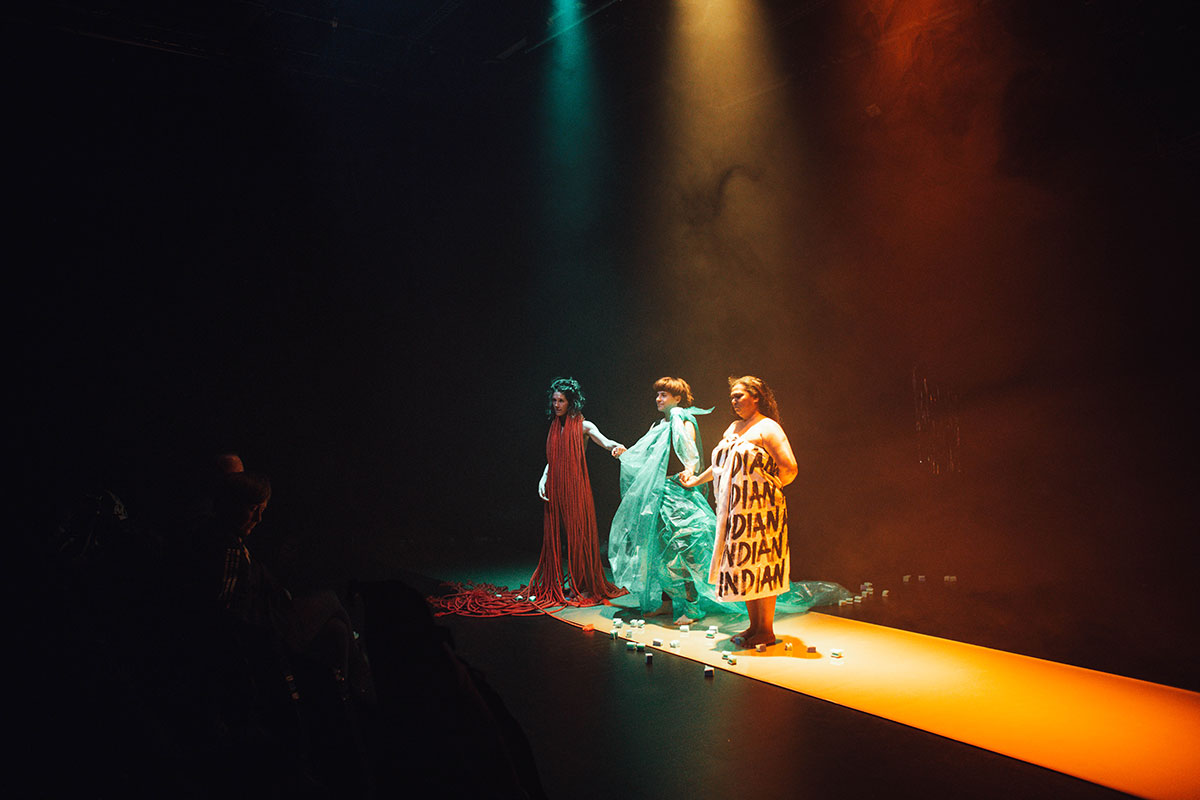 Les trois actrices se tiennent debout sur le plateau. En face du public. C'est le moment des saluts, à la fin du spectacle. La lumière est orange. Nina Langensand, à gauche, est enroulée dans des cordes rouges. Meret Landolt, au milieu, est vêtu de bâches en plastique vertes. Living Smile Vidya, à droite, est enveloppée dans un drap avec marqué plusieurs "INDIAN" dessus.