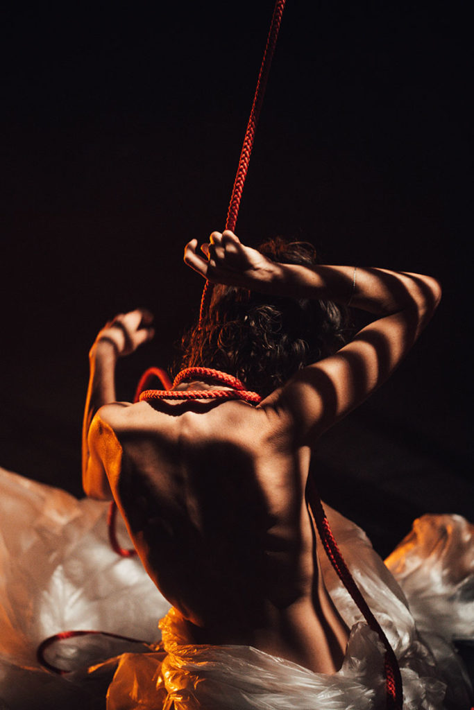 Une des trois actrices est nue, de dos. Elle est assise dans un décor constitué de bâches en plastique blanches. Il y a des contrastes de lumière. Son bras droit est levé pour attraper la corde rouge enroulée autour de son cou.
