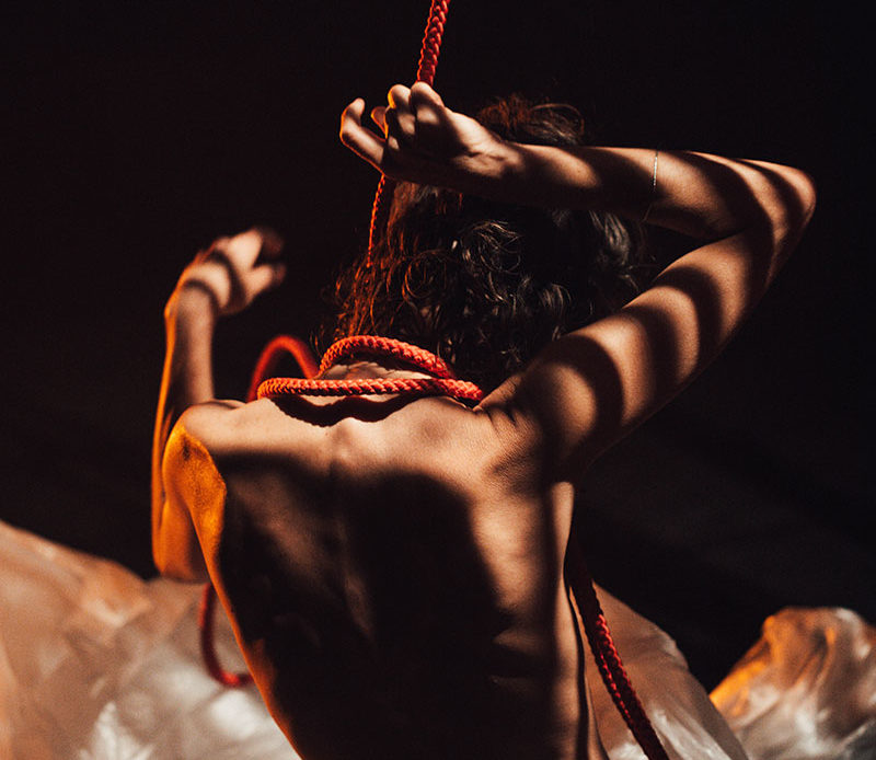 Une des trois actrices est nue, de dos. Elle est assise dans un décor constitué de bâches en plastique blanches. Il y a des contrastes de lumière. Son bras droit est levé pour attraper la corde rouge enroulée autour de son cou.