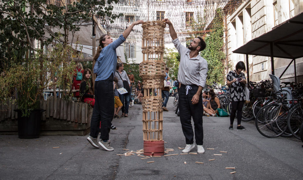 Two people are building a tower in a public space. The tower is made of KAPLA® blocks.