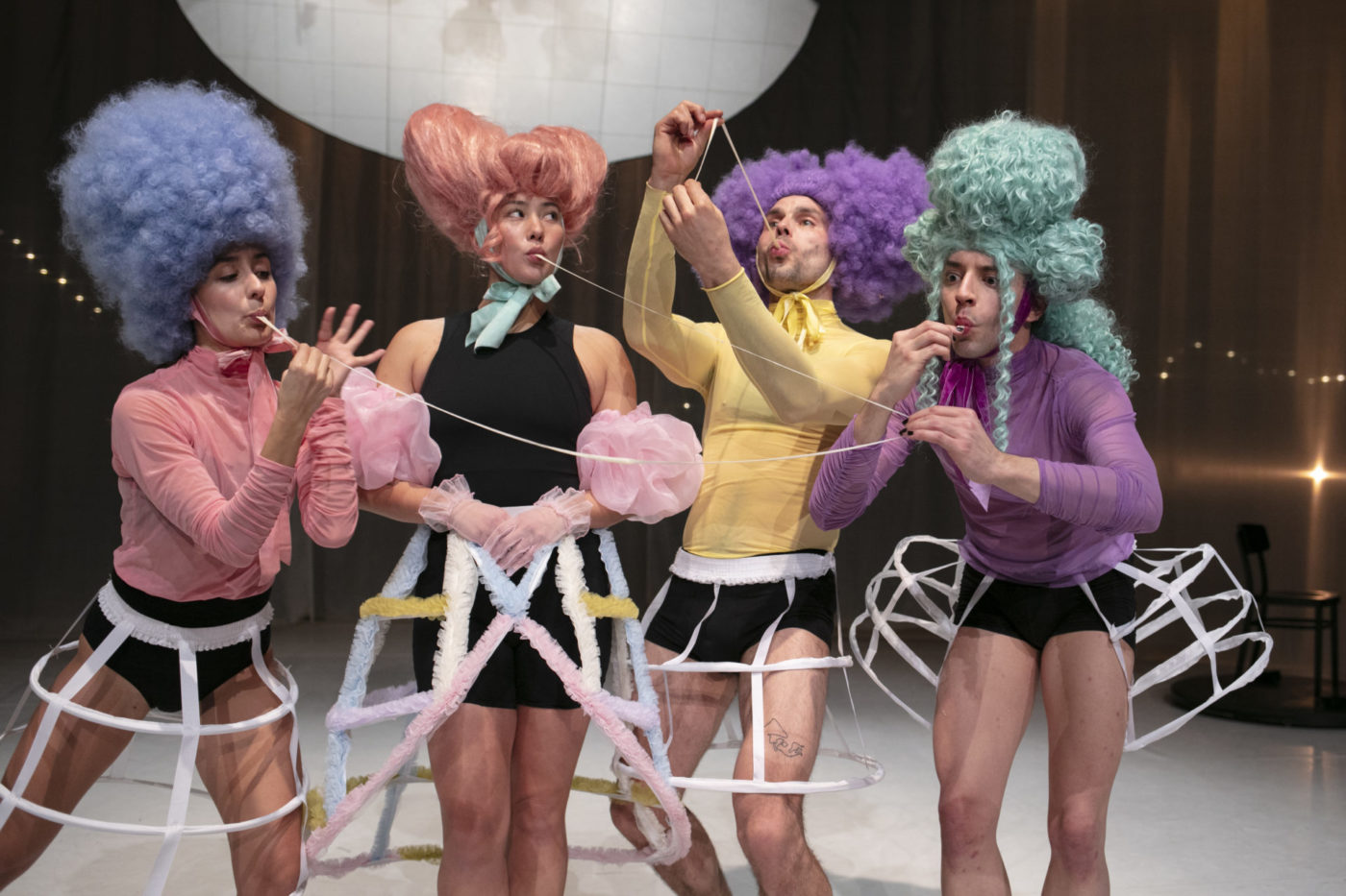 4 danseurs et danseuses sont sur la scène. Ils portent des grandes perruques de différentes couleurs, style 18e. Une bleue, une orange, une violette et une verte. Ils portent des hauts colorés assortis. Ils s'amusent à jouer avec les chewing-gum qui sont dans leur bouche en les étirant. Ils portent une jupe longue quadrillée, qui rappelle ce qu'il y a avait sous les tenues des femmes au 18e siècle.