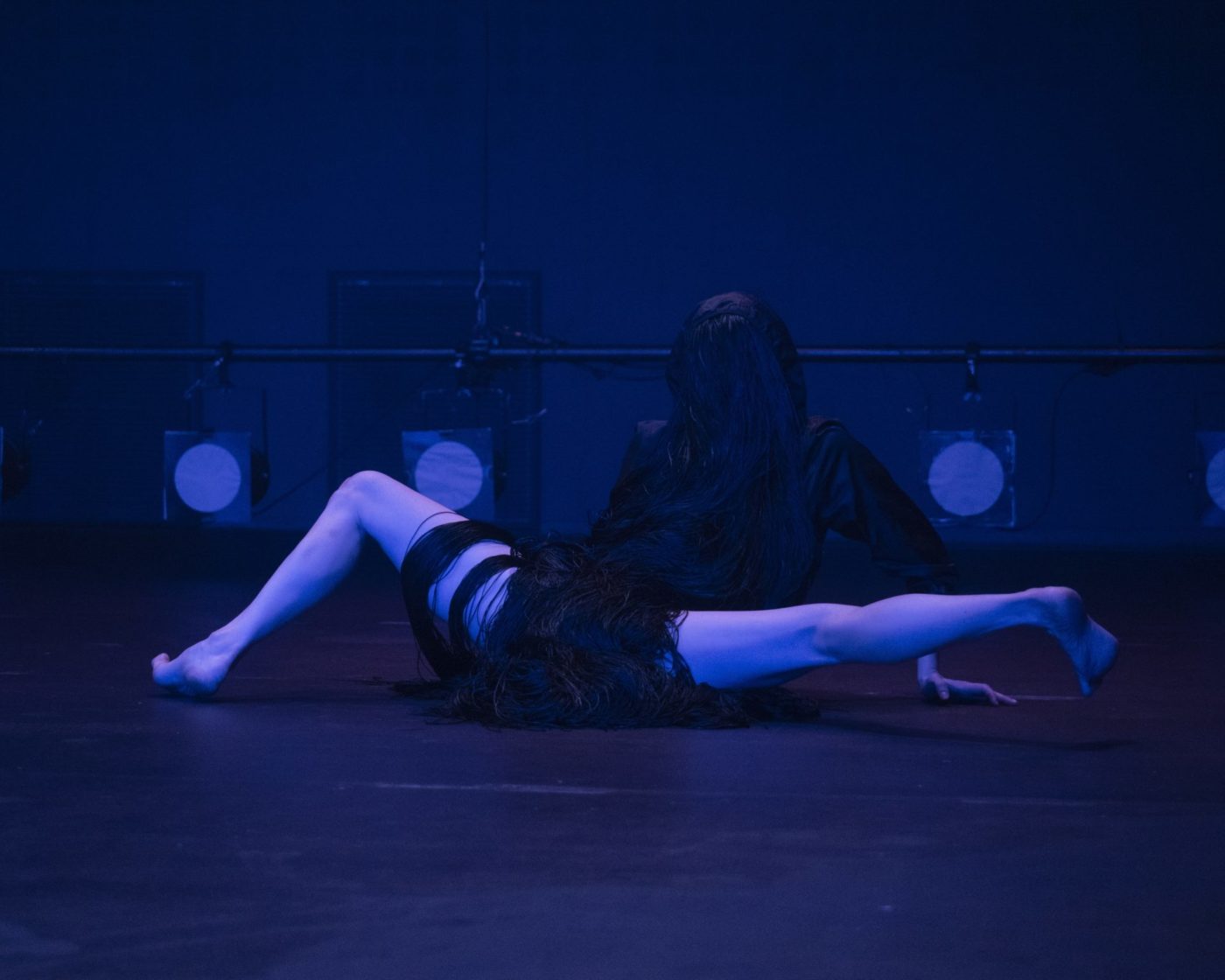 Marion Zurbach est vêtue de noir, dans une salle obscure. Ses jambes sont nues. Elle fait un grand écart. Elle a une perruque noire. Cela donne l'impression d'une forme d'animal étrange.