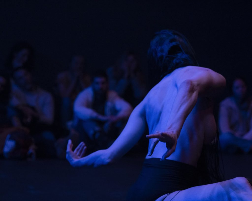 Zoom sur le dos de Marion Zurbach en train de danser. Ses bras sont étendus vers l'arrière de son dos. Le tout est en torsion. La photo est sombre et intrigante.