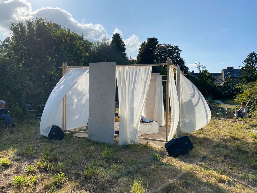 Un lit entouré de rideaux transparents se trouve dehors dans une étendue d'herbe. On aperçoit des personnes à l'intérieur. Les rideaux sont gonflés par le vent. Il fait très beau. Un système son retransmet ce qui se passe à l'intérieur de la structure. Deux personnes écoutent ce qui se passe, assise sur des chaises.