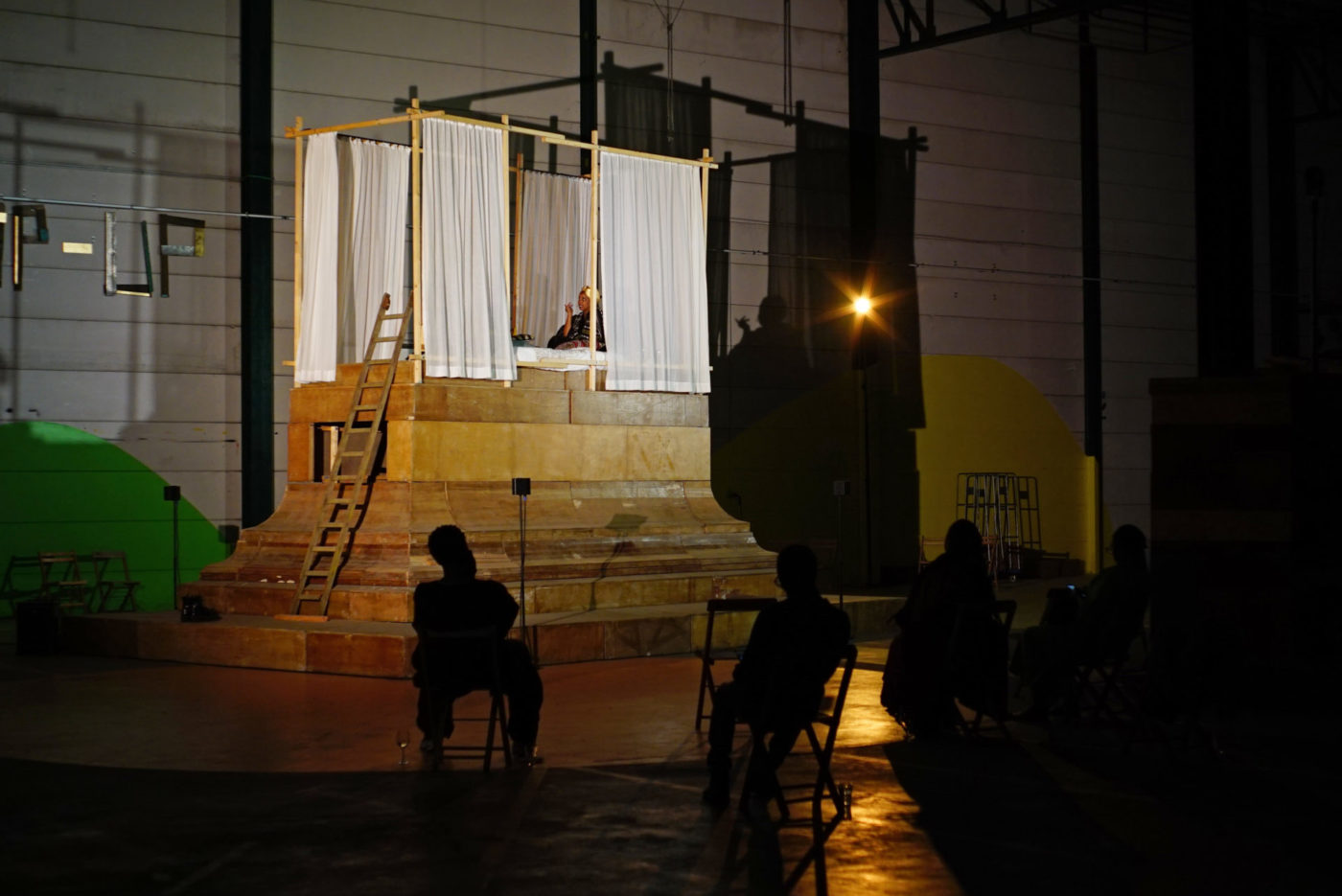 In a theater room, there is an elevated bed bathed in light. The rest of the room is in darkness. The bed is surrounded by curtains. Lucile Saada Choquet is in the bed.