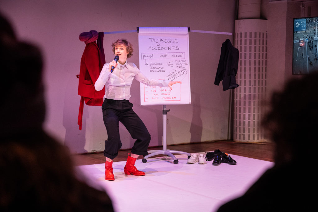Salomé Mooij se tient debout devant un tableau sur lequel elle explique les différentes techniques du trébuchement. Elle porte des bottes rouges et a les genoux pliés, c'est une pause assez marrante. Elle a un micro pour expliquer la situation.