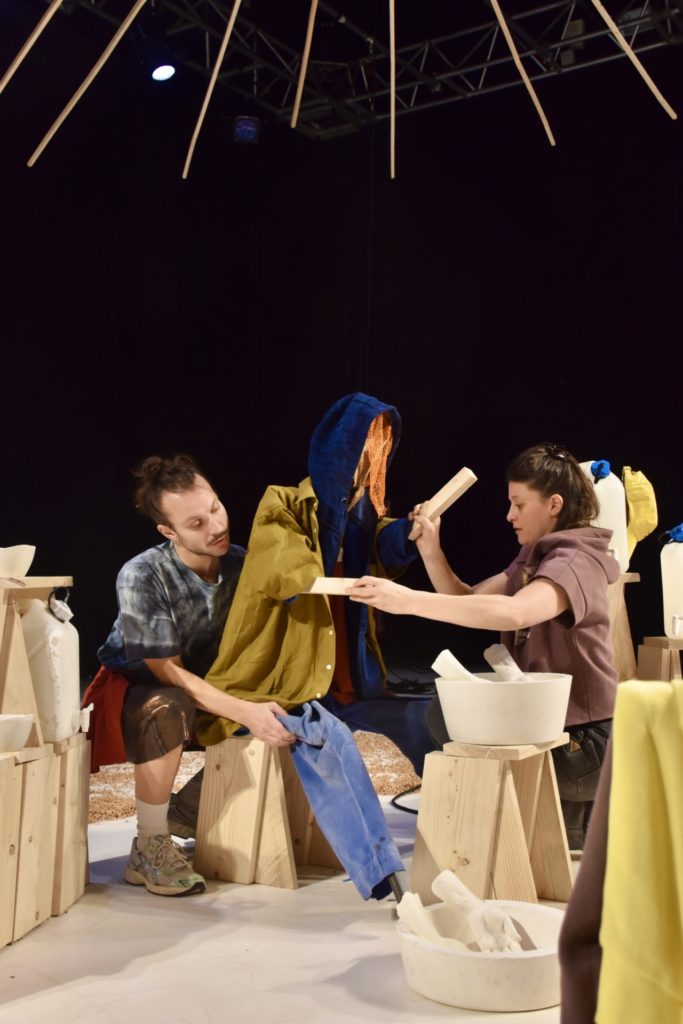 Two performers are dressing a large puppet (life-sized) with human clothes.