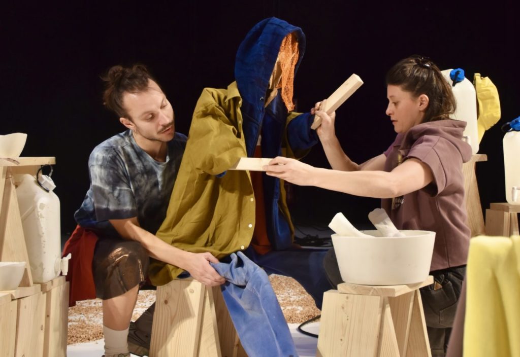 Deux personnes du spectacle sont en train d'habiller une grande marionnette (de taille humaine) avec des vêtements d'humain.
