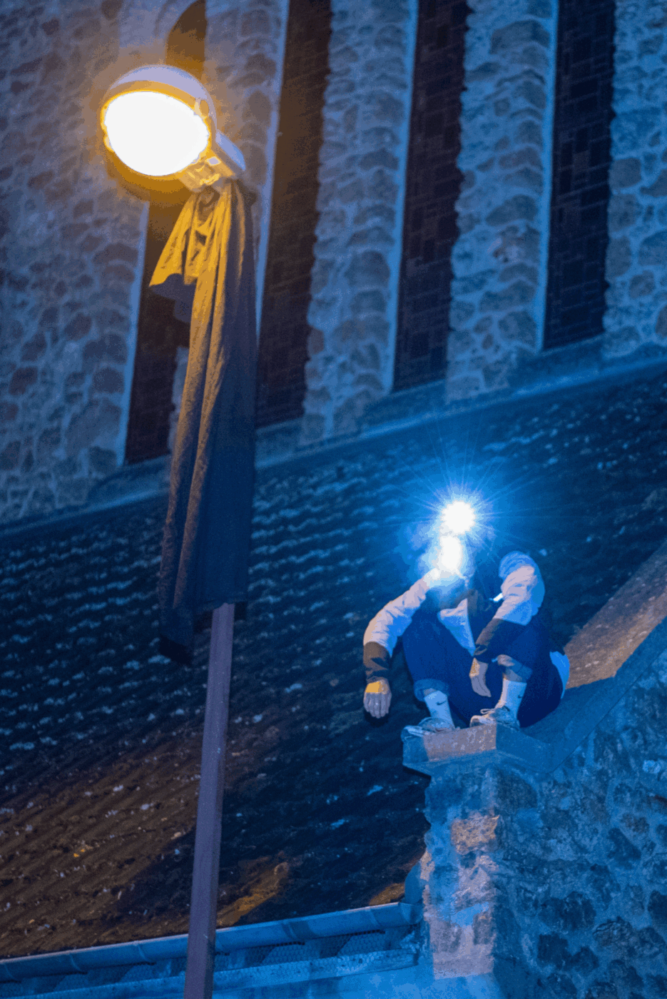 Un homme est assis accroupi sur un toit. Il fait nuit. Il a une lampe frontale. Il y a un lampadaire à côté de lui.