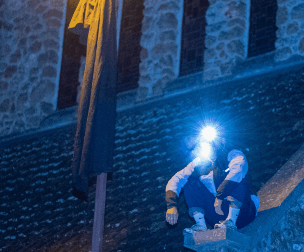 Un homme est assis accroupi sur un toit. Il fait nuit. Il a une lampe frontale. Il y a un lampadaire à côté de lui.