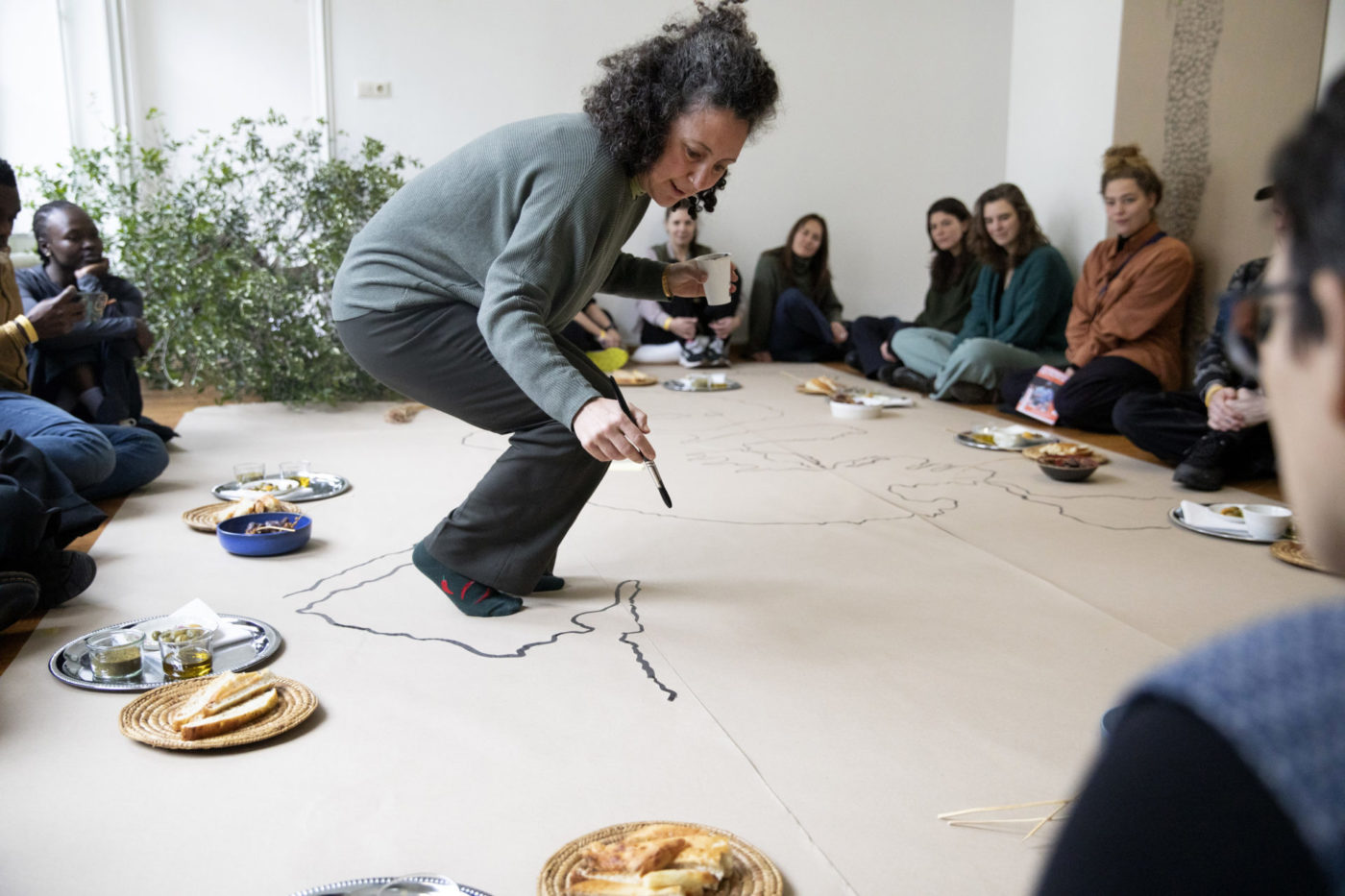 Samah Hijawi se tient debout et dessine une carte au pinceau. La carte est dessiné sur une immense feuille de papier. Du public se tient autour de la feuille. En fond, on aperçoit des branches d'olivier.