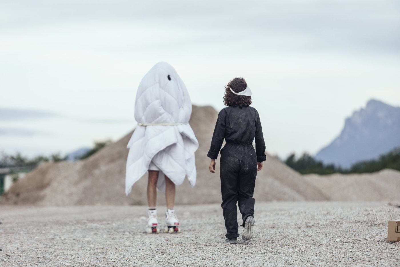 Deux personnes sont en extérieur. Le paysage est un paysages de gravière. Les personnes sont en train de marcher. A droite, la personne est vêtue d'une combinaison noire en tissu. A gauche, la personne est enveloppé dans un grand costume en draps blancs.