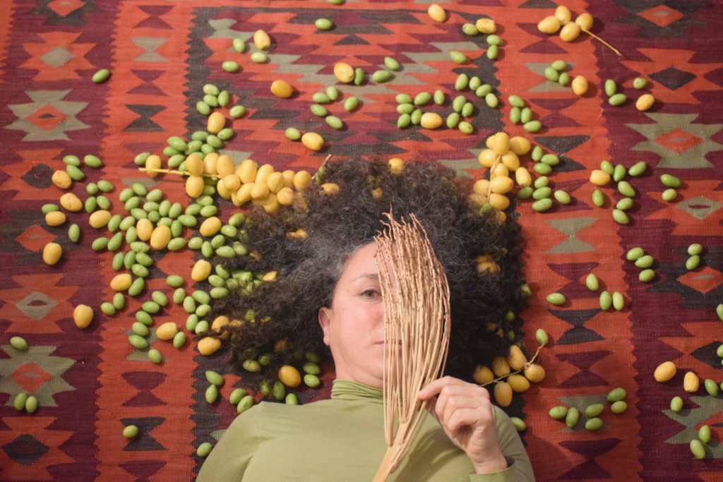 Samah Hijawi est allongée sur un tapis au motif oriental. Il y a des olives jaunes et vertes autour d'elle. Elle brandit une branche sèche d'un arbuste et couvre un de ses yeux avec.