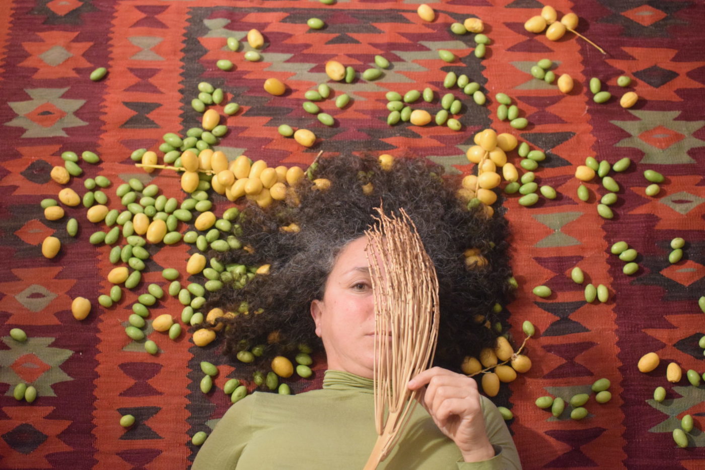 Samah Hijawi est allongée sur un tapis au motif oriental. Il y a des olives jaunes et vertes autour d'elle. Elle brandit une branche sèche d'un arbuste et couvre un de ses yeux avec.