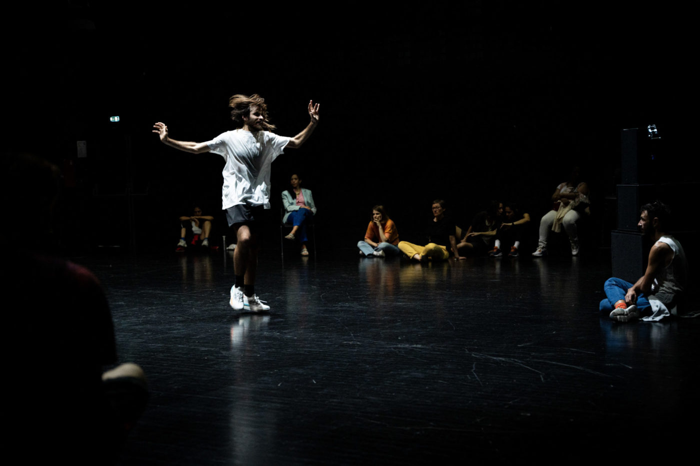 Ametonyo Silva is dancing in a dark room; he looks like he’s bouncing. His hair is tousled, and one knee is raised. The audience is gathered around him in the room.