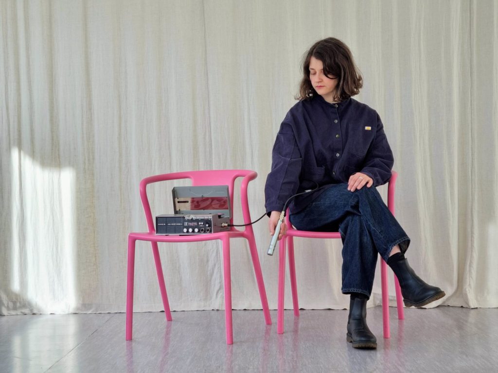 Annina Polivka est assise sur une chaise rouge. Elle tient un micro dans sa main. Le micro est relié à une machine qui produit du son. La machine est également sur une chaise rose, à côté de celle de l'artiste.