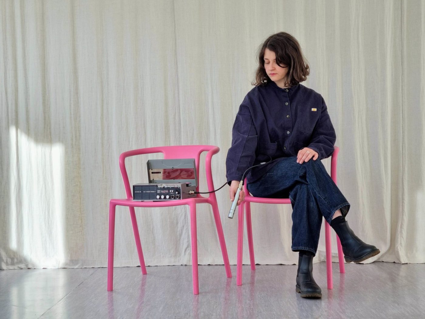 Annina Polivka est assise sur une chaise rouge. Elle tient un micro dans sa main. Le micro est relié à une machine qui produit du son. La machine est également sur une chaise rose, à côté de celle de l'artiste.