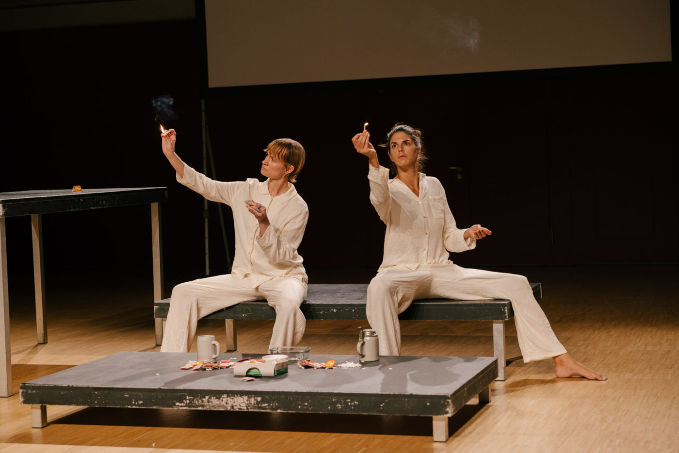 The two artists Flavia Papadaniel and Diane Dormet sit on a bank on the scene. They wear pyjamas. Each of them holds a lighted match in the right hand, looking at it fixedly.