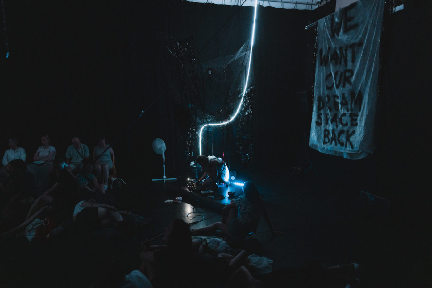 The room is only enlighted thanks to some blue neon lights. We can see the artist Francesca Sproccati crouched on her music keyboard. She produces sounds. On her left we discover a banderole on which it is written "We want our dream space back".